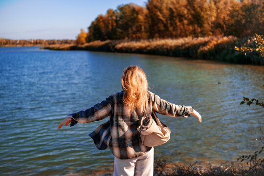 Relaxed woman by autumn lake spreading her arms &mdash; mental health connection. Female traveler standing on shore breathing fresh air enjoying landscape. Digital detox and natural mindfulness concept.