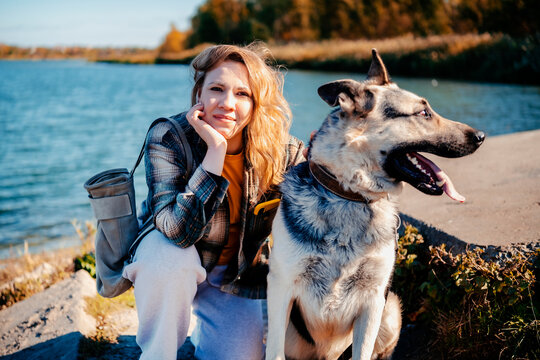 Cheerful young woman squatting beside east european shepherd dog near river. Urban pet friendly environment and active lifestyle concept. Woman cynologist enjoying sunny autumn day with animal.