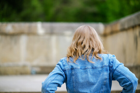 Woman from behind in denim jacket outdoors near stone wall in park. Casual style concept for outdoor travel, rear view portrait, and autumn fashion.