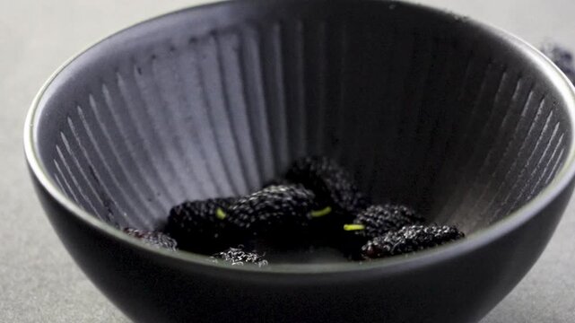 Ripe black mulberry fruits falling into black ceramic bowl on dark background, 240fps