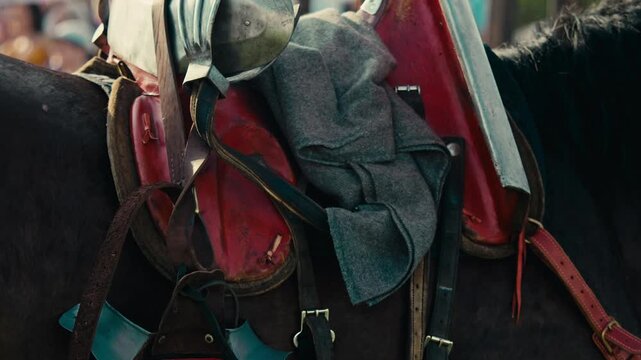 Close up reveals red medieval saddle with steel armor on horse. Grey blanket and leather straps secure heavy riding equipment for upcoming historical jousting tournament.
