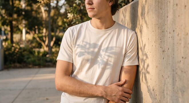 Medium close-up of a young man wearing a blank off-white crewneck t-shirt outdoors against a concrete wall with natural sunlight shadows.