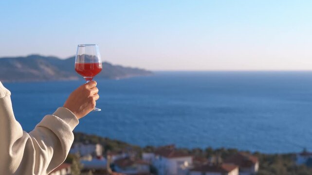 Woman holding glass of rose wine enjoying sea view. Woman's hand holding a glass of rose wine, gently swirling it against a beautiful, sunny backdrop of the blue sea and a coastal town