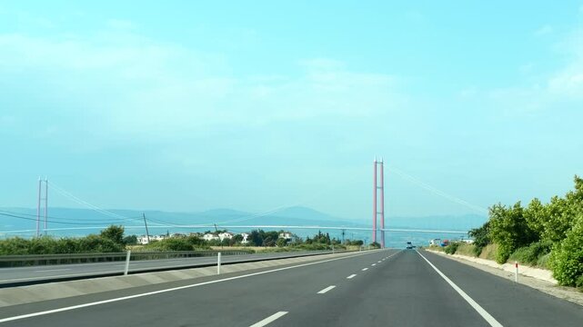 Car driving along a highway approaches a large suspension bridge spanning over water. A sunny day features blue skies and fluffy clouds, enhancing the scenic journey