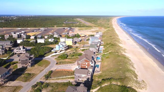 Beachfront homes border grassy dunes beside a long shoreline and breaking ocean waves.
