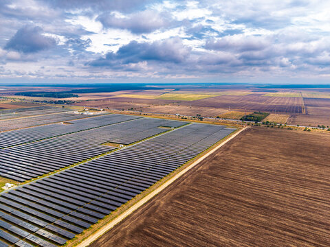 Solar panel installation bordering a brown agricultural field. Long module rows meet an unpaved perimeter road beneath layered clouds and a flat horizon.