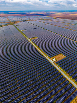 Solar power plant, aerial perspective of photovoltaic arrays across terrain. Numerous rows of dark modules cover vast level ground under scattered white clouds and bright daylight.