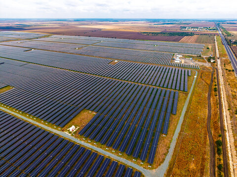 Solar farm beside a straight road and electrical equipment compound. Parallel module rows spread across open fields, with pale utility structures near the right edge.