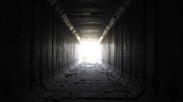 Dark unfinished concrete subway tunnel with repeating structural segments and bright daylight at the end.