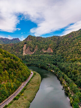 River valley, aerial overview of winding waterway and coastal road. Dense green vegetation blankets steep hills alongside a calm flowing stream and transit route.