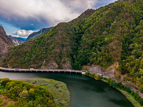Riverside viaduct curving below a wooded mountain slope. Vehicles cross a road supported by pillars above dark water, with reeds and trees along the near bank.