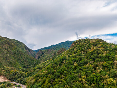 Forested mountain ridges beneath a low cloud layer. Green and yellow trees cover steep hills, with exposed rock near a road at the lower left edge.