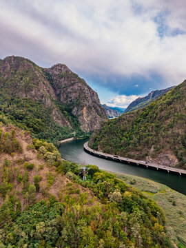 Mountain gorge, aerial shot of winding river and bridge with vehicles. Steep rocky slopes covered in dense green forest enclose a serpentine water channel and viaduct.