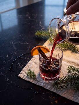 Hand pouring warm aromatic mulled wine from a glass carafe into a crystal tumbler garnished with cinnamon and orange slice on a festive table