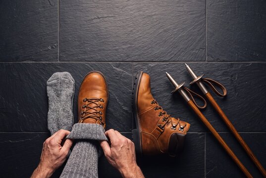 High angle view of a hiker putting on warm wool socks and leather boots next to trekking poles on a dark stone floor