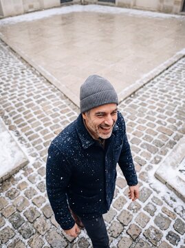 Happy mature man in knit beanie and warm coat enjoying snowfall outdoors on a cobblestone street
