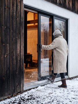 Person in Warm Winter Coat Shaking Snow Off Boots Before Entering Cozy Modern Cabin Through Sliding Glass Door