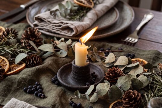 Rustic holiday table setting with burning candle, eucalyptus leaves, pinecones, dried orange slices, and ceramic tableware