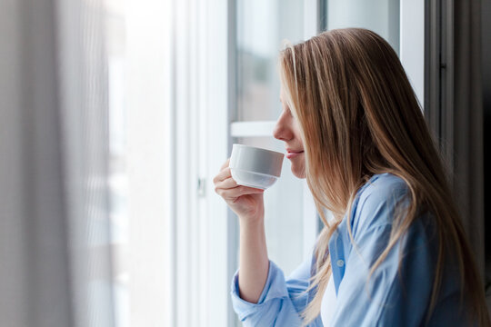 Young woman drinking coffee in hotel room, enjoying morning routine, calming drink. Solo traveler relaxing by opened window. Slow travel, mindfulness, female mental health. Happy girl in vacation