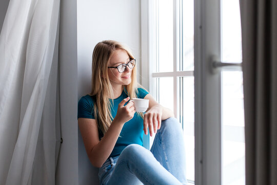 Young woman relaxing on windowsill, looking out opened window. Airing out room, ventilation. Happy girl enjoying morning coffee, calming drink at home. Slow living, mindfulness, female mental health