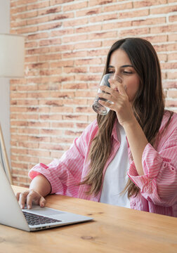 Young woman working from her home office, focused on her laptop while taking a refreshing sip of water to stay hydrated and maintain wellness during a productive workday