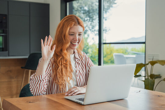 Smiling young redhead woman working remotely, communicating with colleagues or friends during a video conference, enjoying modern technology and flexible workspace from home