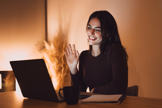 Young woman smiling and waving at her laptop screen during a video call, communicating remotely from a home office at night, showcasing modern virtual connection and remote work lifestyle