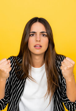 Young woman expressing strong negative emotions, clenching her fists in frustration and anger, standing against a vibrant yellow background, conveying intense emotional stress and disappointment