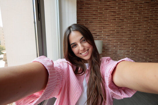 Young woman smiling broadly, stretching arms out, and capturing a selfie indoors, showing a happy and confident lifestyle moment in natural light against a brick wall and window