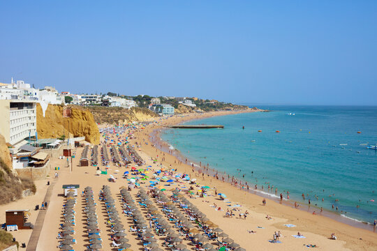 Aerial view of Albufeira, Algarve, Portugal, showing a busy sandy beach, turquoise Atlantic Ocean, colorful umbrellas, coastal cliffs, and whitewashed buildings under a clear blue sky.
