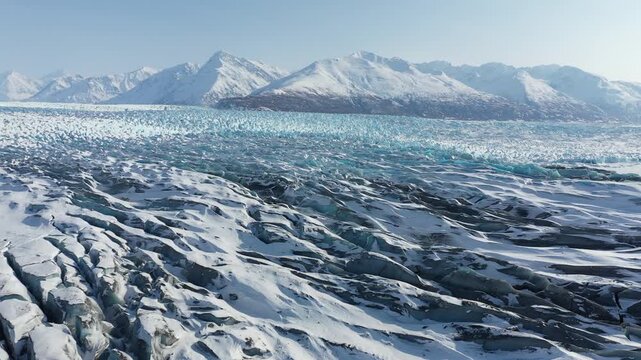 Drone Low Pass Over Textured Blue Glacial Ice Surface and Crevasses with Snowy Peaks on the horizon