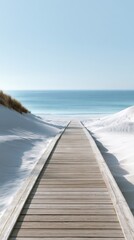Fototapeta premium Wooden boardwalk leading through snowy dunes toward the calm ocean under clear blue sky on a bright winter day