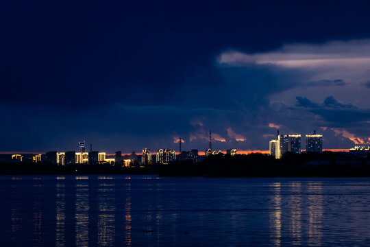 Dramatic dusk city view across Zeya river, capturing vivid tower lights, dark clouds and glowing horizon
