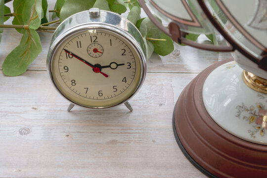 Close-up view of a vintage alarm clock and an elegant floral table lamp on a white wooden table with eucalyptus. Perfect for cozy bedroom lifestyle, time management, and night routine backgrounds.