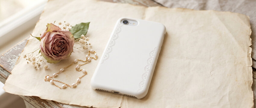 White smartphone with lace-trimmed case beside a rose and pearl necklace on handmade paper. Blank smartphone mockup for app design. Mobile branding and product presentation. Copy space