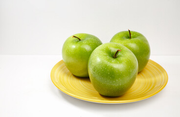 Three fresh green apples on a yellow plate against a clean white background, representing healthy eating, nutrition, freshness and wholesome lifestyle. © Anastasia.photoyoga