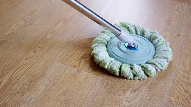 Up-close shot of a microfiber spin mop head moving across a gleaming laminate wood floor, leaving behind a damp trail. Reflecting the theme of household cleaning and daily tasks