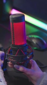 Hand with purple nail polish adjusts red and black microphone on desk revealing details like mesh grille and shock mount. background features colorful LED lighting and computer peripherals