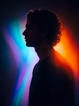 Silhouette of a young man with curly hair illuminated by vibrant neon blue and orange lighting in studio