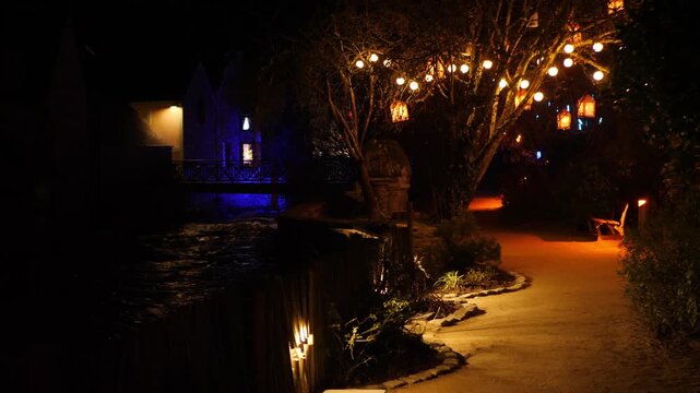 Serene night illuminated walkway beside dark water. Bare trees strung with warm orange accent lights create festive ambiance halloween and christmas. Bluelit building structure visible left.