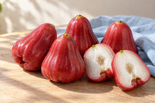 Red wax apples and lengthwise halves reveal crisp white flesh and hollow centers on a light wooden table with pale blue fabric.
