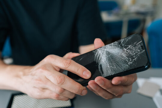 Hands examining damaged mobile device with shattered glass display. Concept of concepts of device repair, accidental breakage, need for screen replacement, close-up, cropped shot
