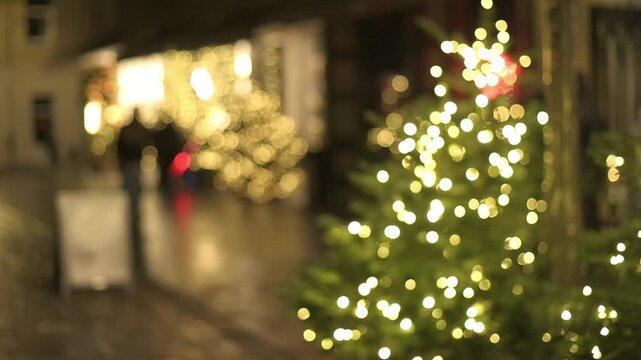 Street view featuring glowing Christmas trees and blurred figures of shoppers strolling by festive storefronts in a lively urban environment