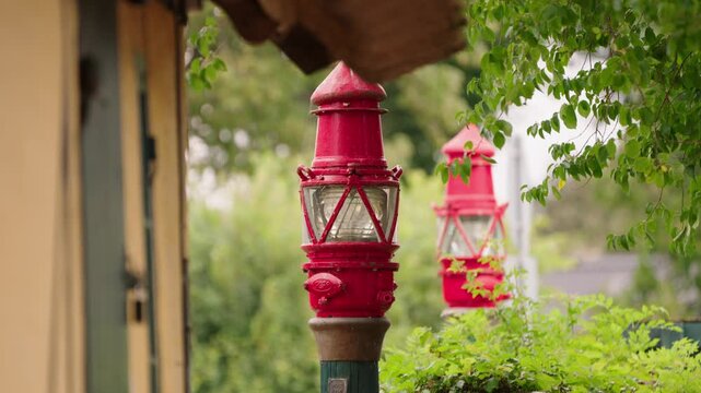 Close view of two vintage red maritime signal lanterns mounted outdoors, showing weathered metal, glass lenses and traditional nautical design among green foliage.