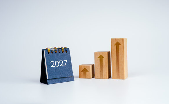 2027 desk calendar beside rising wooden blocks as graph steps with upward arrows. Business growth, financial performance, annual planning, investment outlook and strategic goals for the coming year.
