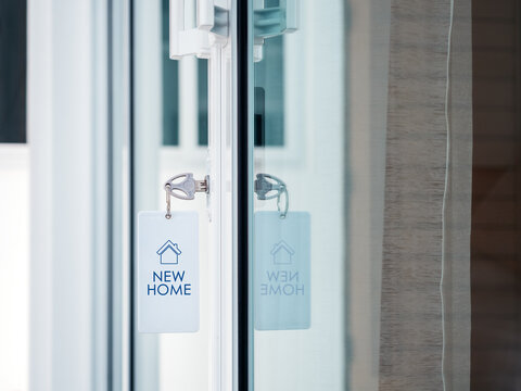 Key with new home tag hangs from modern residential glass sliding door. Homeownership, property purchase, moving in, secure access and new beginning in real estate concept.