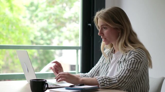A young blonde woman diligently works on her laptop from home. She takes a thoughtful pause, looking out the window, perhaps pondering an idea or momentarily distracted from her tasks.