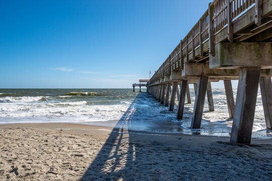 A wooden pier extends over rolling surf from a sandy beach on a clear coastal day.