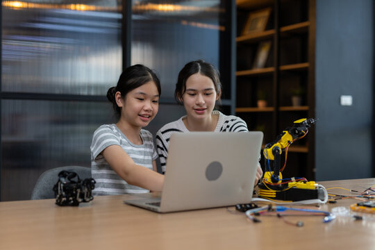Happy Asian girl and woman smiling while learning robotics programming on laptop computer together with small robotic arm on desk