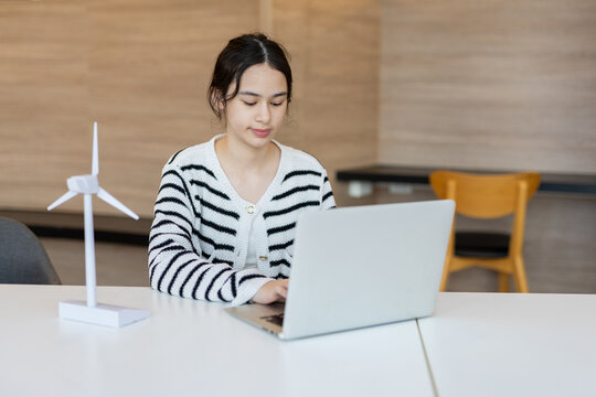 young asian woman student using laptop computer at desk with model wind turbine studying renewable energy education